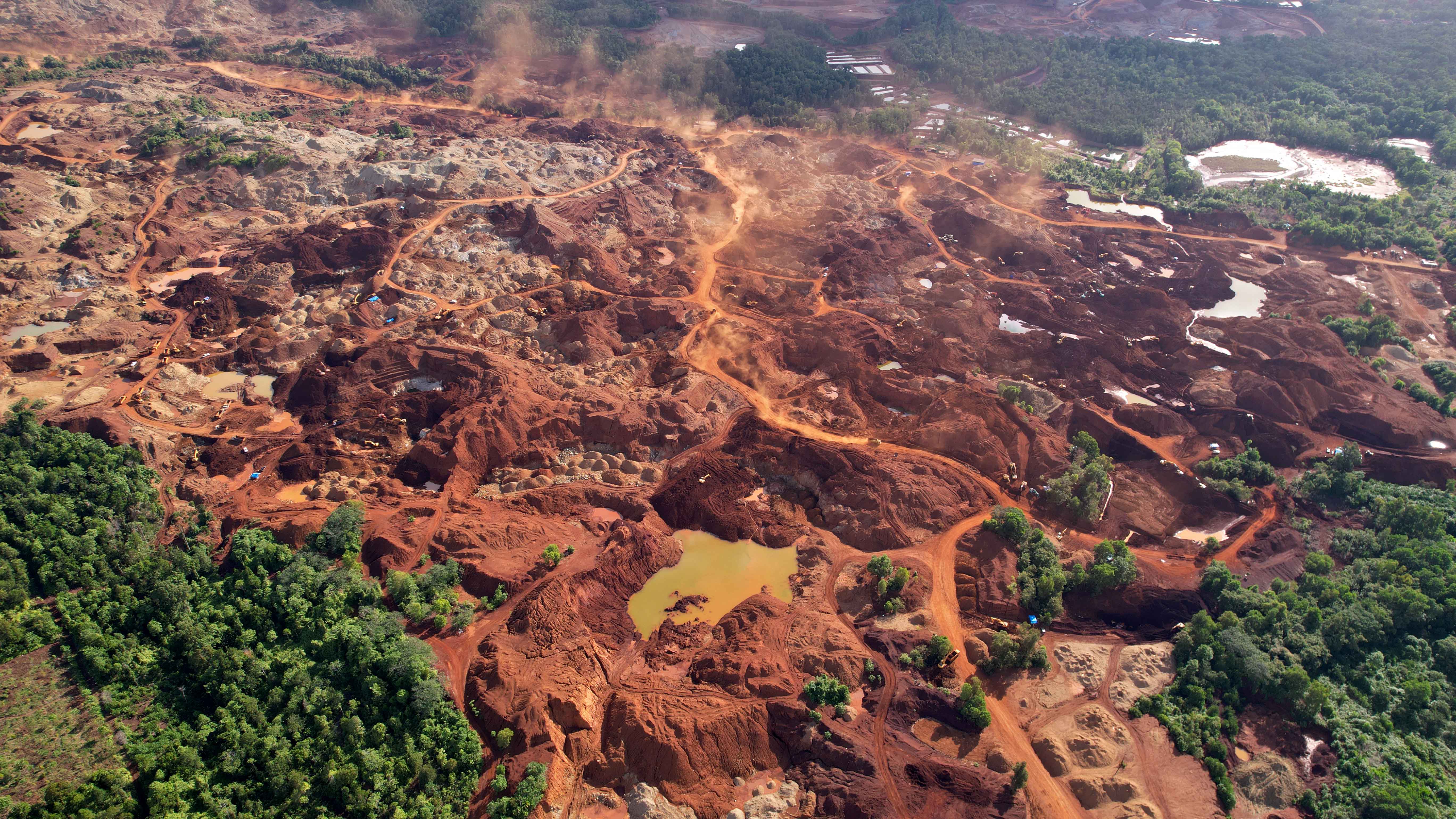 An aerial view of mining landscape with yellow pools of water and dust rising from behind trucks driving through.