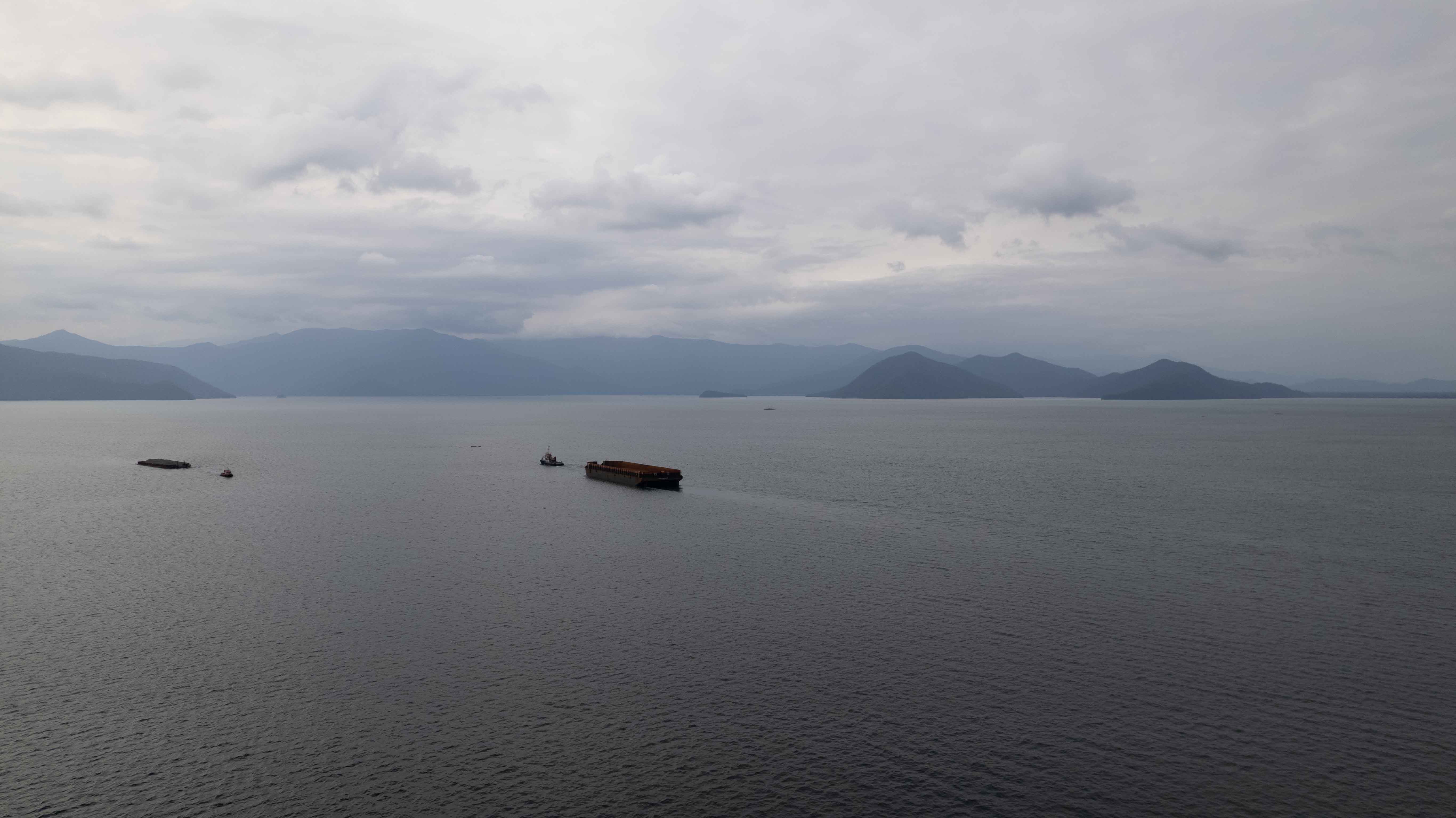 A tug boat pulls a large barge behind it through the sea with the mountainous shoreline in the distance.