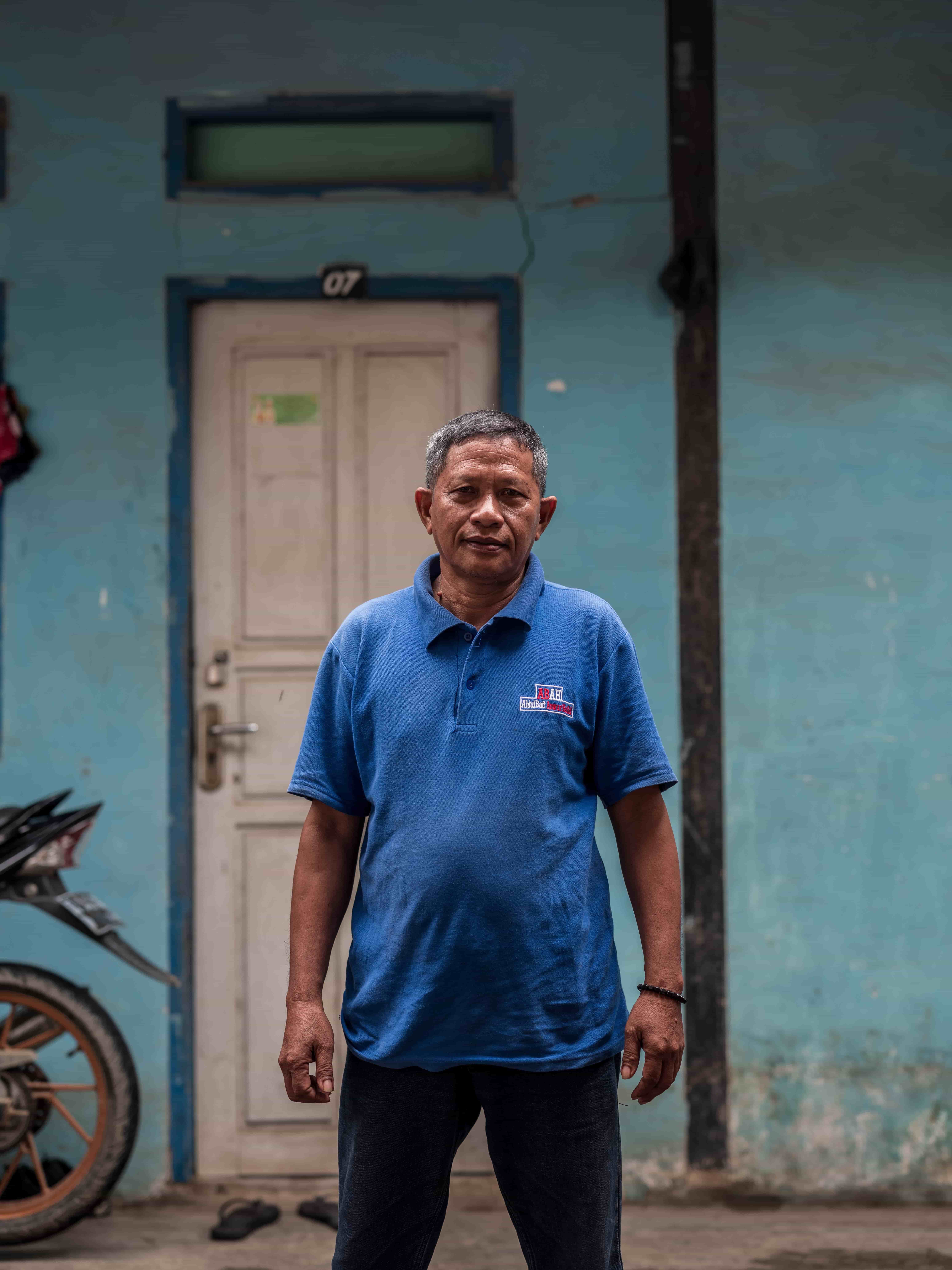 A man in a short-sleeved blue polo shirt poses for a portrait with a turquoise wall and white door behind him.