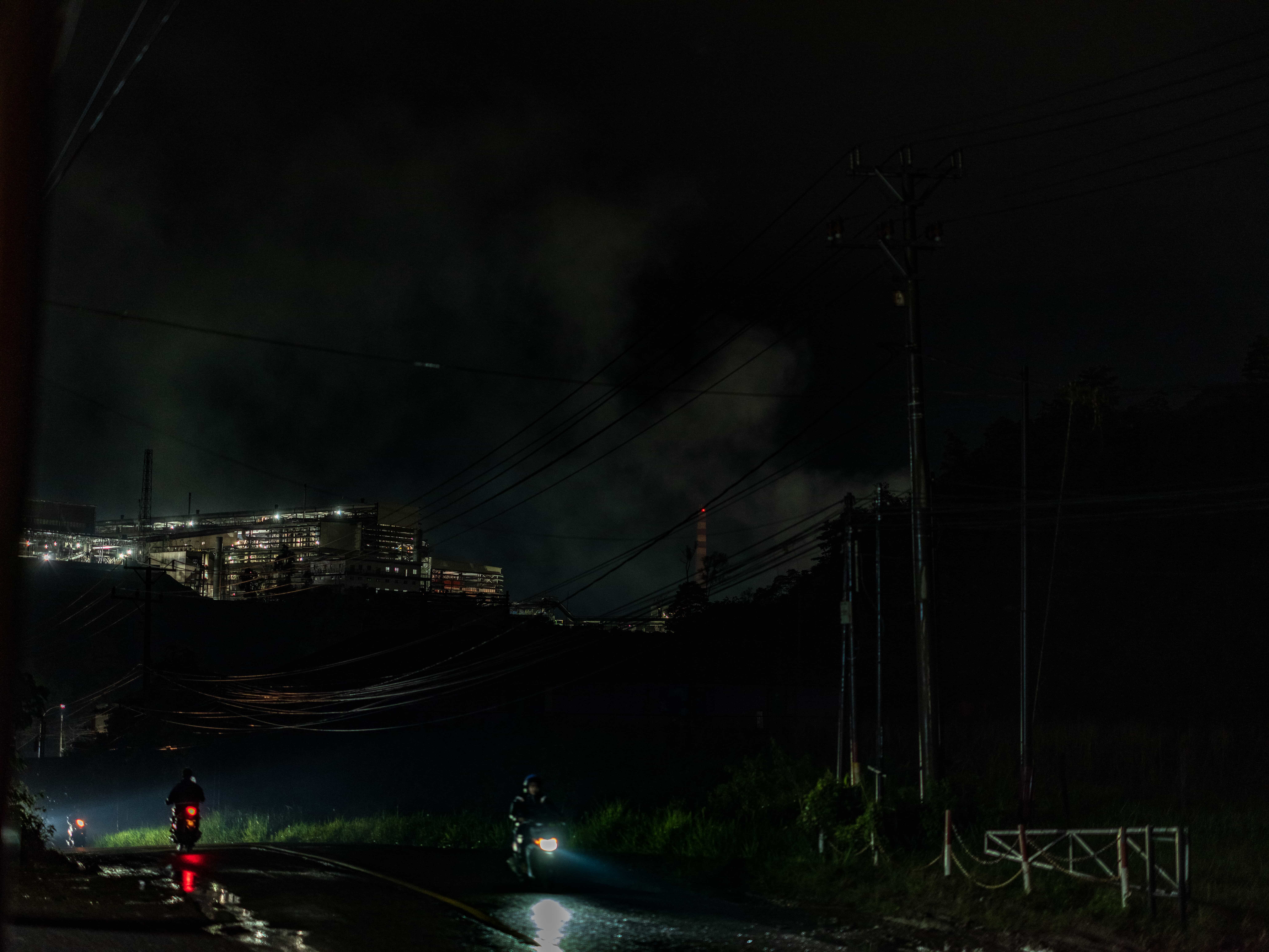 A moped driver rides at night on a dark road with a mining facility lit up behind him on a hill.