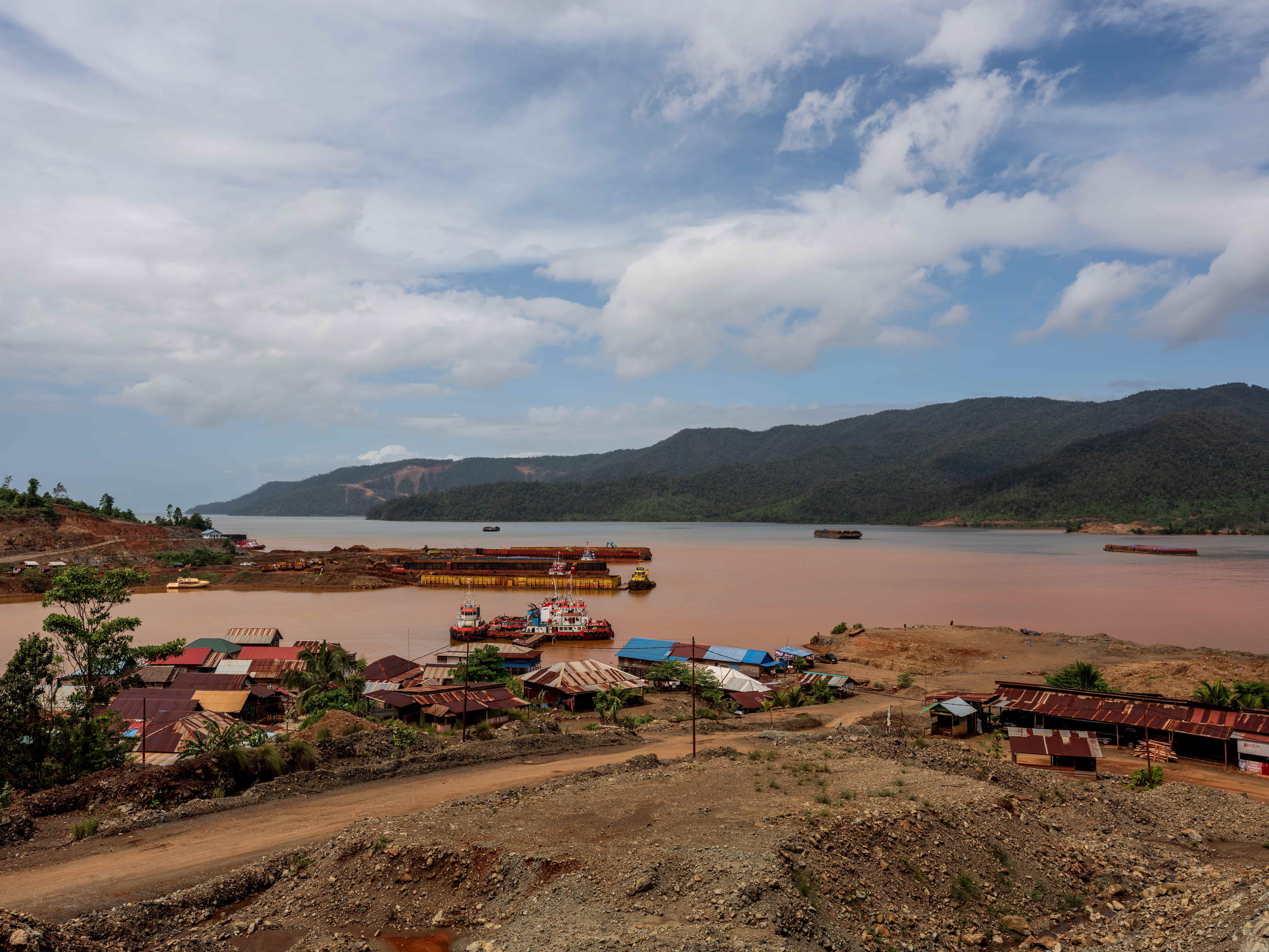 A muddy bay filled with boats and a jetty for mining transportation. A dirt road cuts past a number of small houses with rusting metal roofs.