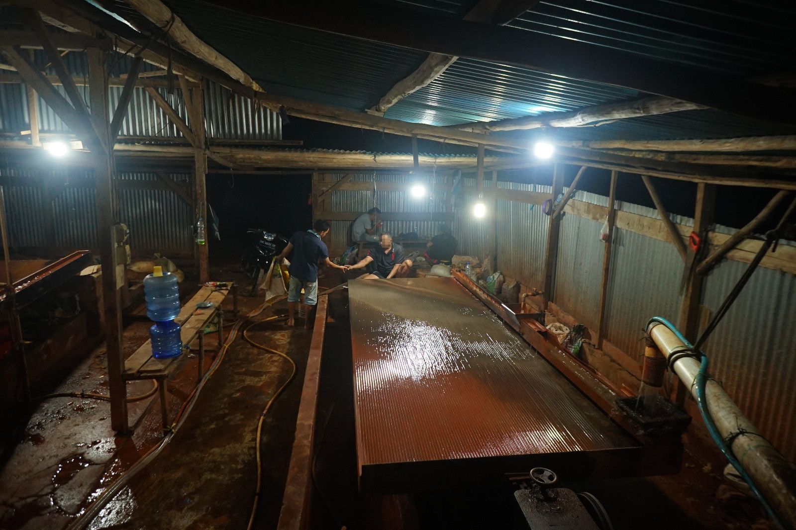 Several men stand behind a large shiny brown metal table in a dark warehouse.