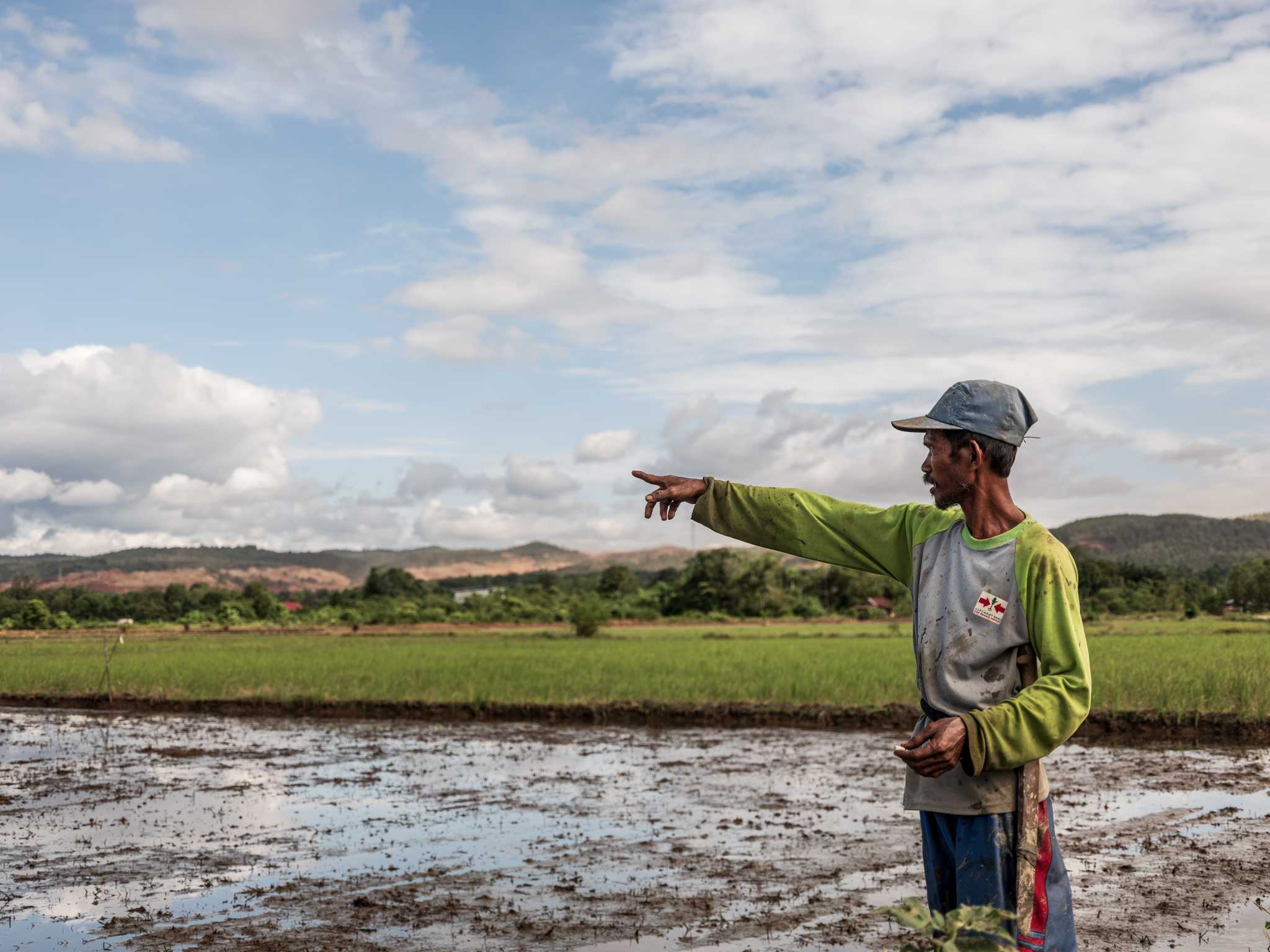 A rice farmer stands in his muddy field looking and pointing away to the left.