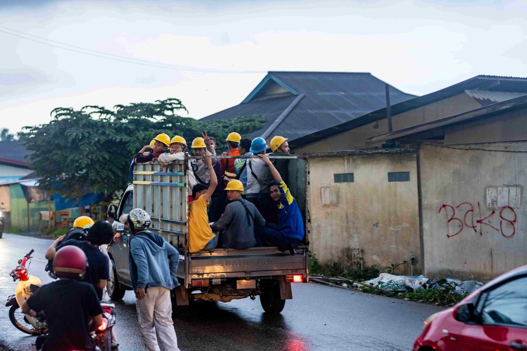 Dozens of miners in yellow helmets ride in different directions on moped scooters at a busy intersection near a mining facility.