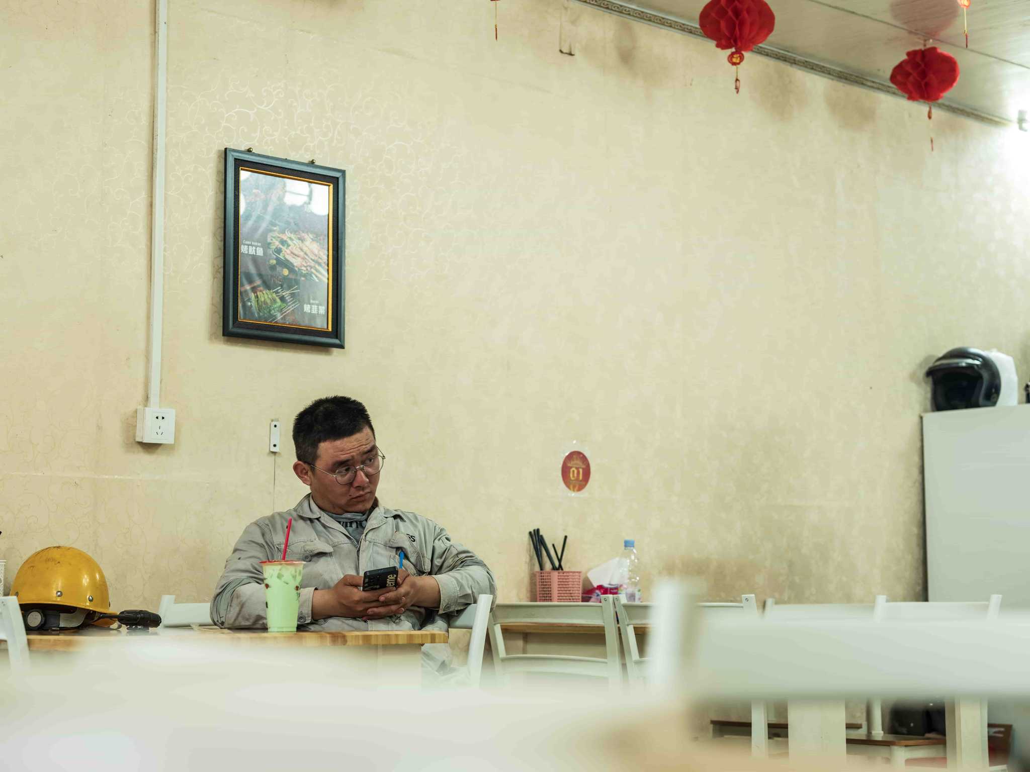 A Chinese worker sits at a table with his yellow work helmet laying on the table and red paper lanterns hanging from the ceiling.