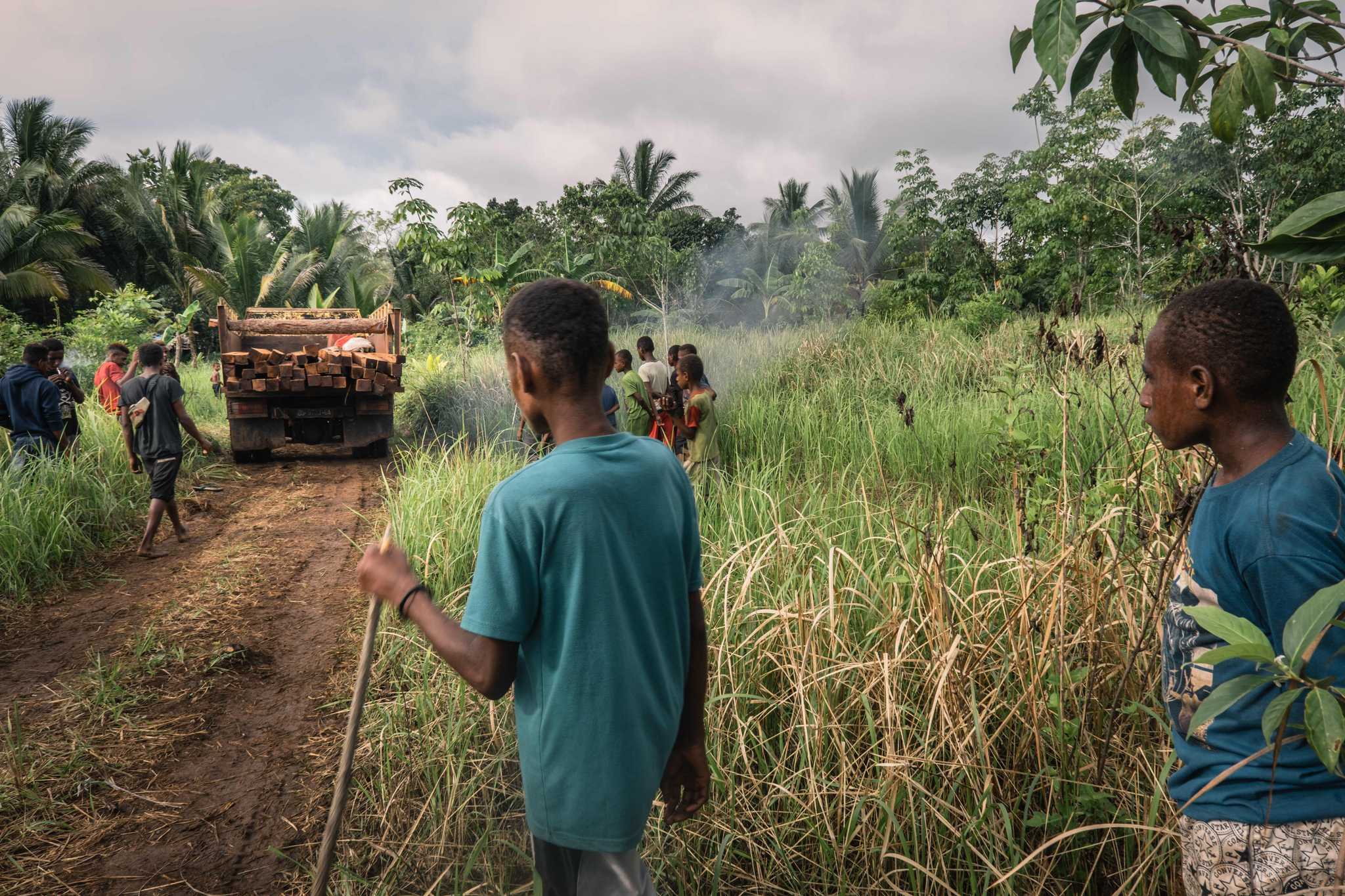 Laki-laki dan anak-anak menonton sebuah truk yang membawa kayu tebangan melintas di jalan menuju kampung Zanegi.